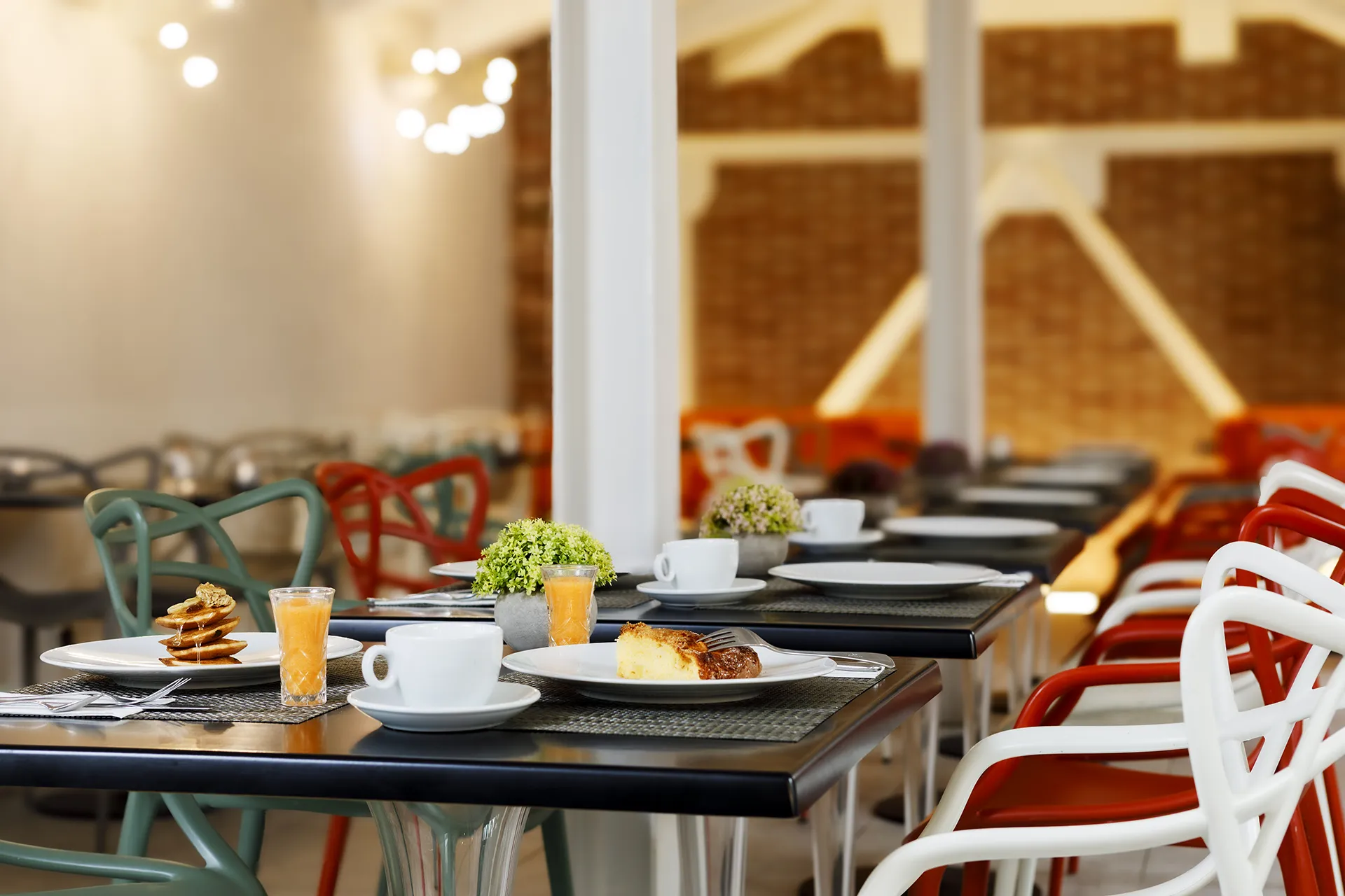 Tavolo apparecchiato per la colazione in un ambiente moderno, con piatti contenenti pancake e una fetta di torta, tazze di caffè, bicchieri di succo d’arancia e piccoli centrotavola floreali. Sullo sfondo si vedono altre sedie colorate e tavoli disposti in fila, in una sala luminosa con pareti chiare e lampadine decorative sospese.